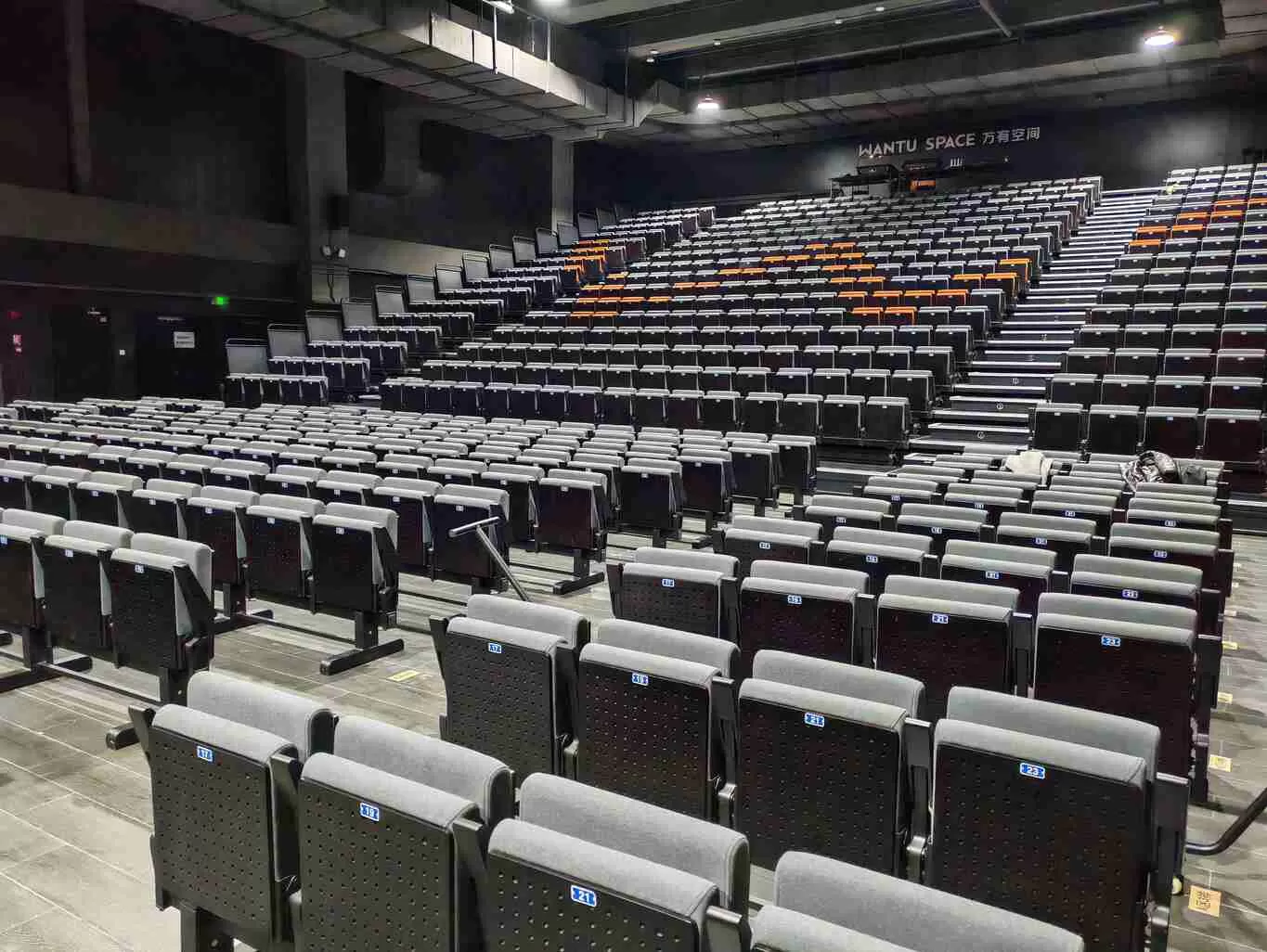 Interior of WANTU SPACE theater: Rows of gray-and-black auditorium seats with blue number labels, some orange-accented seats in the background, and a staff member standing on the right